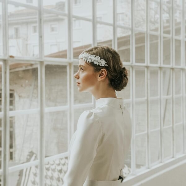 Pensive young female in white elegant dress and hairband standing near windows in room in daylight