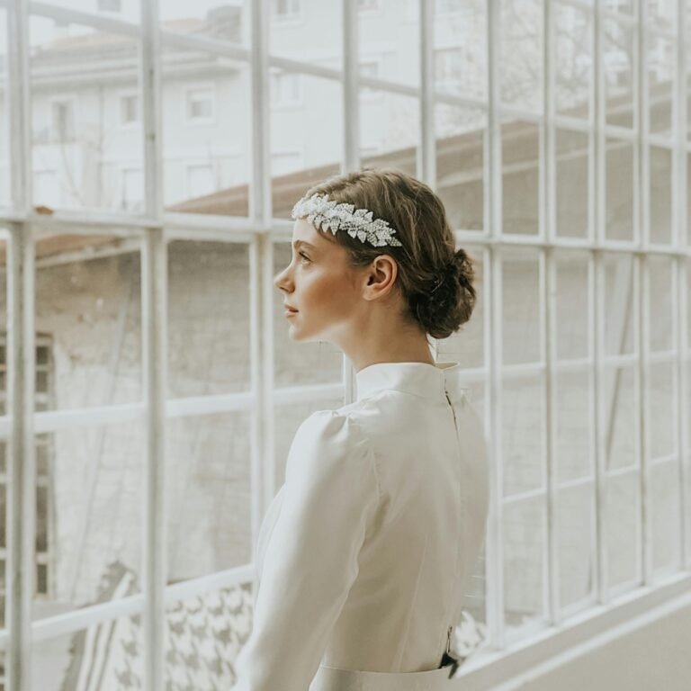 Pensive young female in white elegant dress and hairband standing near windows in room in daylight