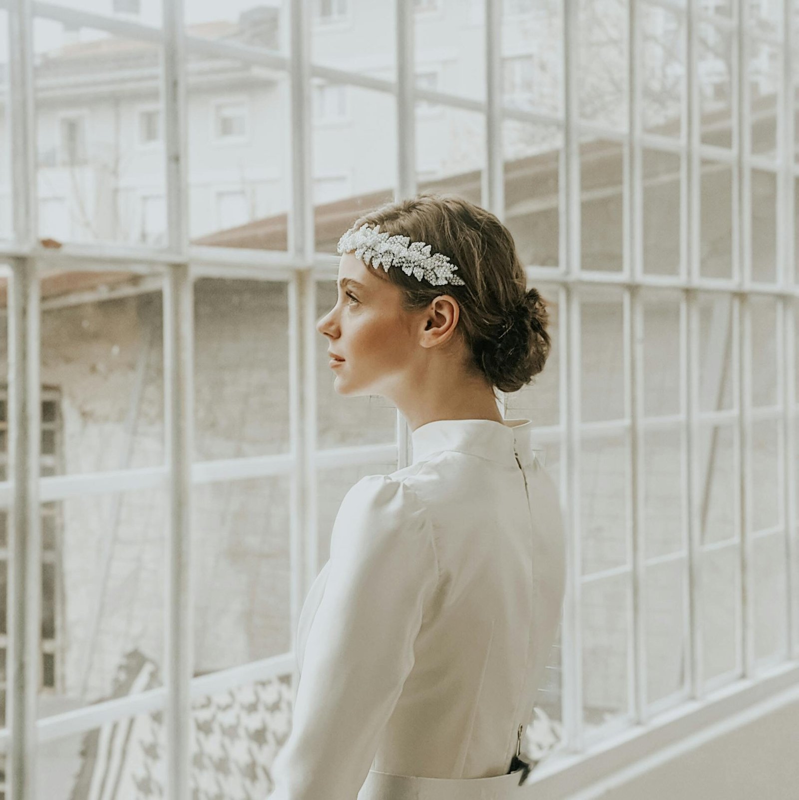 Pensive young female in white elegant dress and hairband standing near windows in room in daylight