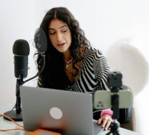A young woman using a microphone and laptop to record a podcast indoors, showcasing a modern content creation setup.