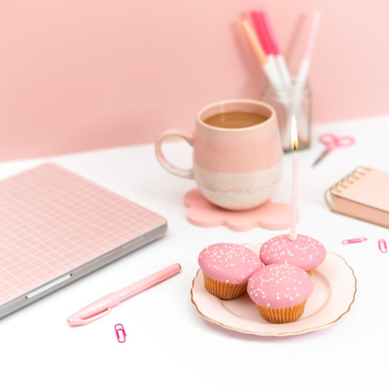 Elegant pink-themed workspace featuring a laptop, cupcakes with candles, and coffee mug.