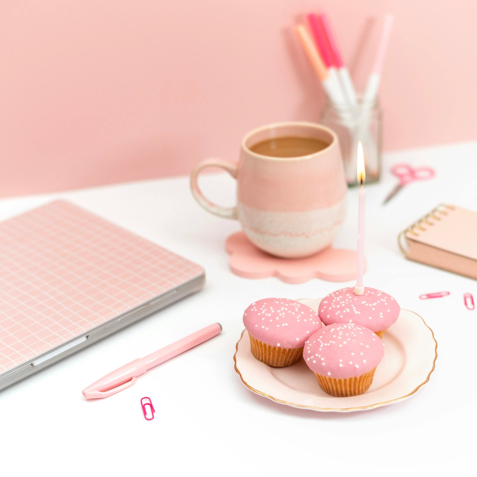 Elegant pink-themed workspace featuring a laptop, cupcakes with candles, and coffee mug.