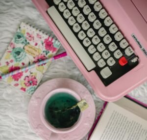 Flat lay of a vintage pink typewriter, open book, and tea on a cozy workspace.