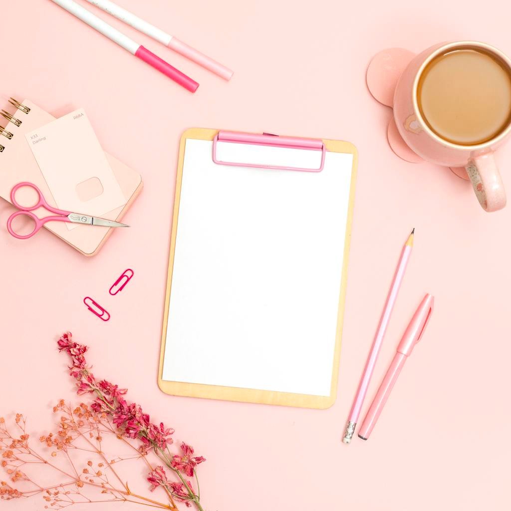 Minimalist flat lay of pink office supplies featuring a clipboard, stationery, and coffee cup.