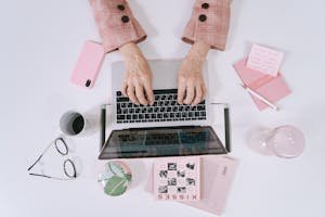 Stylish overhead shot featuring hands typing on a laptop with pink office accessories for a chic workspace vibe.