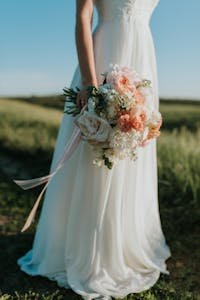 A bride in a flowing white gown holds a colorful bouquet in a sunny field.
