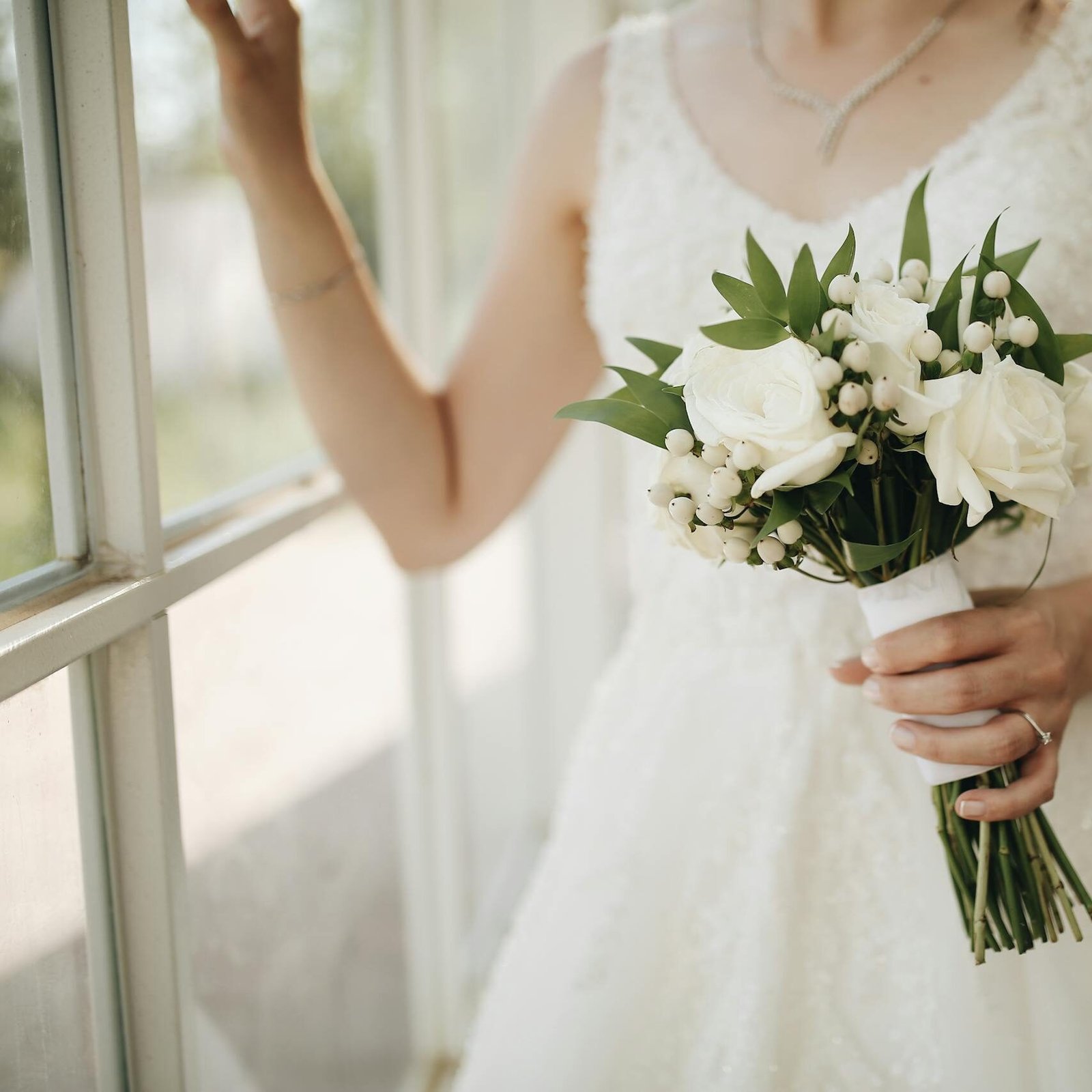 Bride in a white dress holding a rose bouquet near a window, capturing elegance and romance.