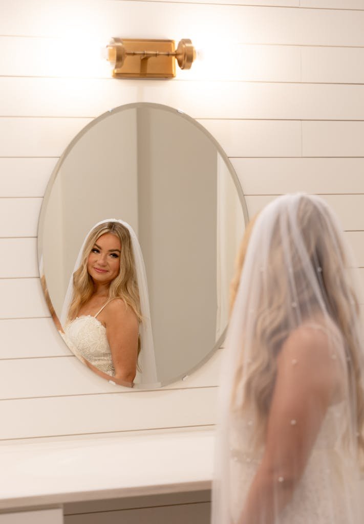 Elegant bride in a white wedding dress looking into a round mirror indoors.