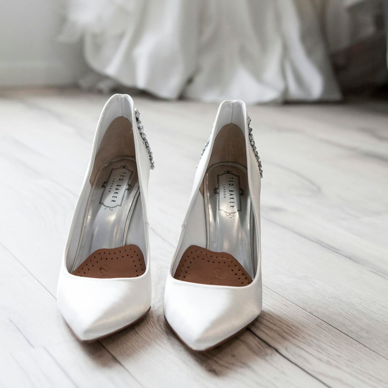 Stylish white bridal shoes positioned in front of a flowing wedding dress indoors.